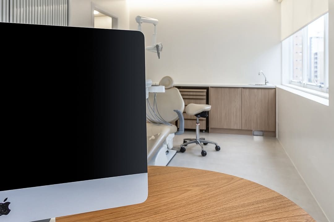 Consultório com mesa de madeira e computador, e a área de atendimento ao fundo