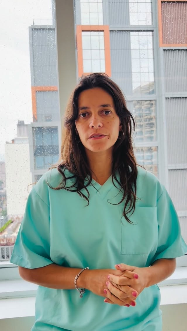 Dra. Joana Ozi, com scrubs verde-água, na clínica em frente à janela com vista para os prédios de São Paulo